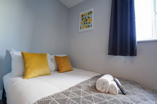 Una habitación de hotel con una cama, dos almohadas, una colcha estampada, una toalla enrollada, un control remoto y un cuadro en la pared.