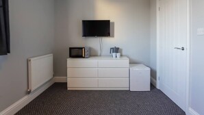 Una habitación pequeña y funcional con un aparador blanco, un microondas, un refrigerador compacto, un televisor de pantalla plana montado en la pared y una puerta blanca.