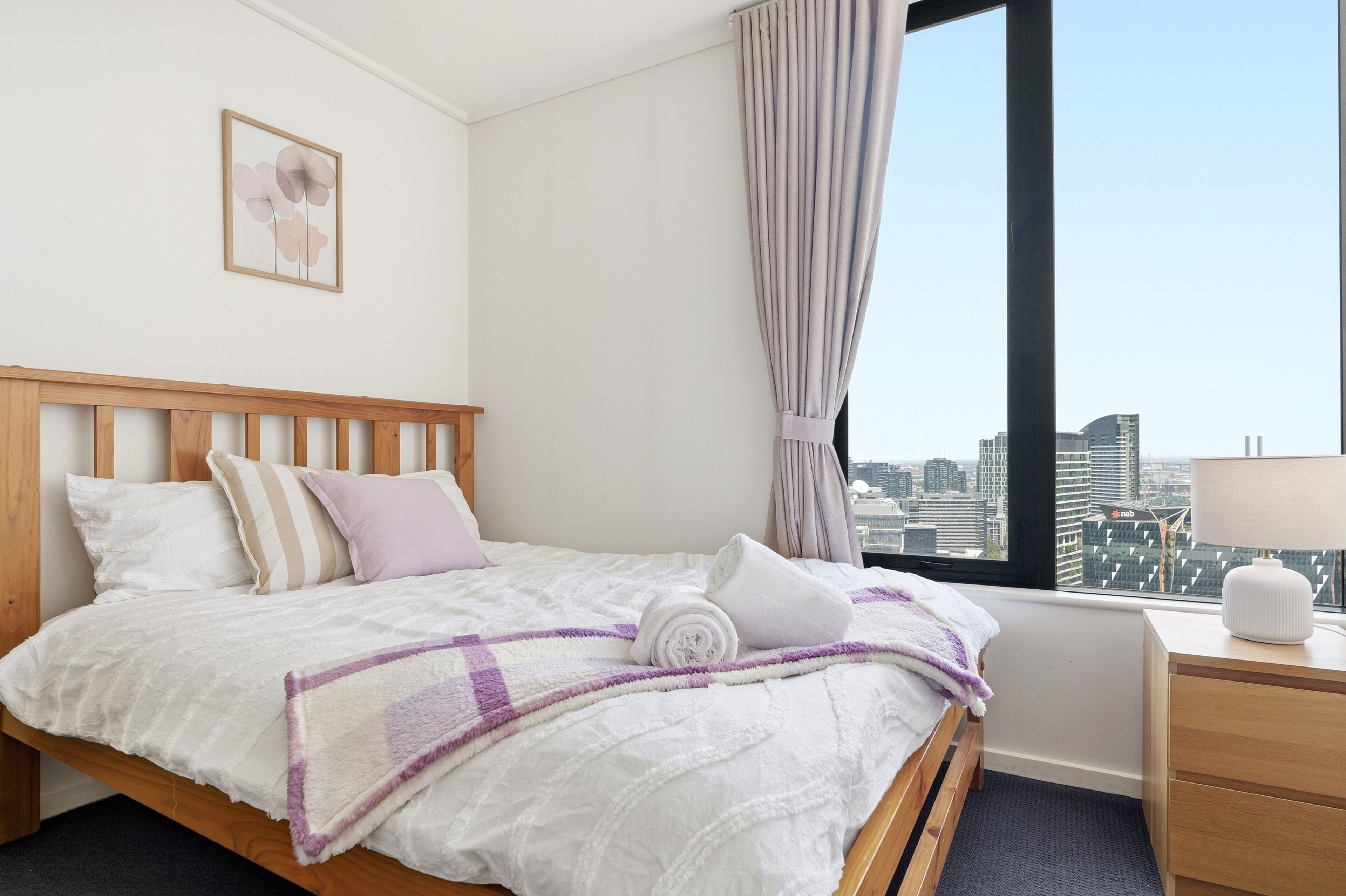A hotel room with a bed, a nightstand, a lamp, a window with a city view, and a framed picture on the wall.