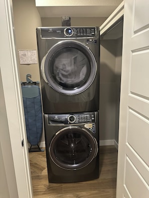 A stacked washer and dryer unit in a laundry room with a blue ironing board and a hanging garment. - Spacious 2-bedroom Basement Suite Near West Edmonton Mall. (Edmonton)