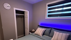 Un dormitorio con cama, mesita de noche, reloj, ventana con persianas e iluminación en el techo.