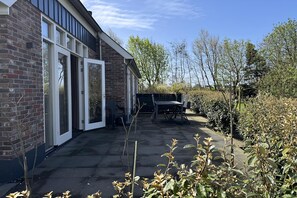 Ein Backsteinhaus mit großen Glasfenstern, einer Terrasse mit Gartenmöbeln und umgebender Bepflanzung.