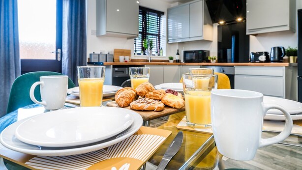 Ein Frühstücksaufstellplatz mit Tellern, Tassen und Gebäck auf einem Tisch in einer modernen Küche mit Schränken und einem Fenster.