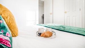 Ein ordentlich bezogenes Bett mit Tablett, auf dem Croissants, Kaffee und Beeren liegen, eine grüne Tagesdecke und eine weiße Wand mit einer Tür im Hintergrund.