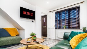 Ein Wohnzimmer mit einer Couch, einem Couchtisch mit Snacks, einem Fernseher, der 'NETFLIX' anzeigt, und einem Fenster mit Jalousien.