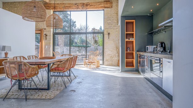 Una cocina moderna con horno integrado y microondas, un comedor con sillas de mimbre y una mesa de madera, y ventanas amplias que ofrecen vistas al exterior.