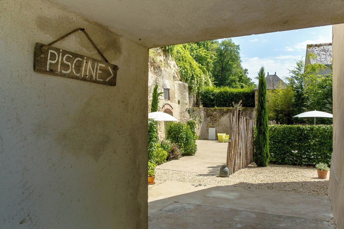 Un espacio al aire libre rústico con un cartel que dice 'PISCINE', un muro de piedra y un sendero de grava que lleva a un edificio.