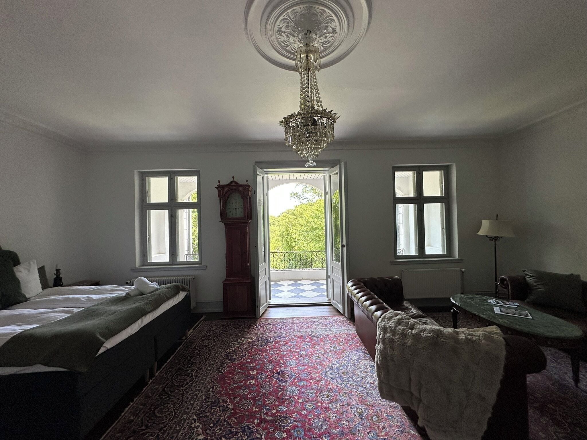 Ein Zimmer mit einem Bett, einer Bank, einem Tisch, einer Chandelüster und Blick durch eine geöffnete Tür.