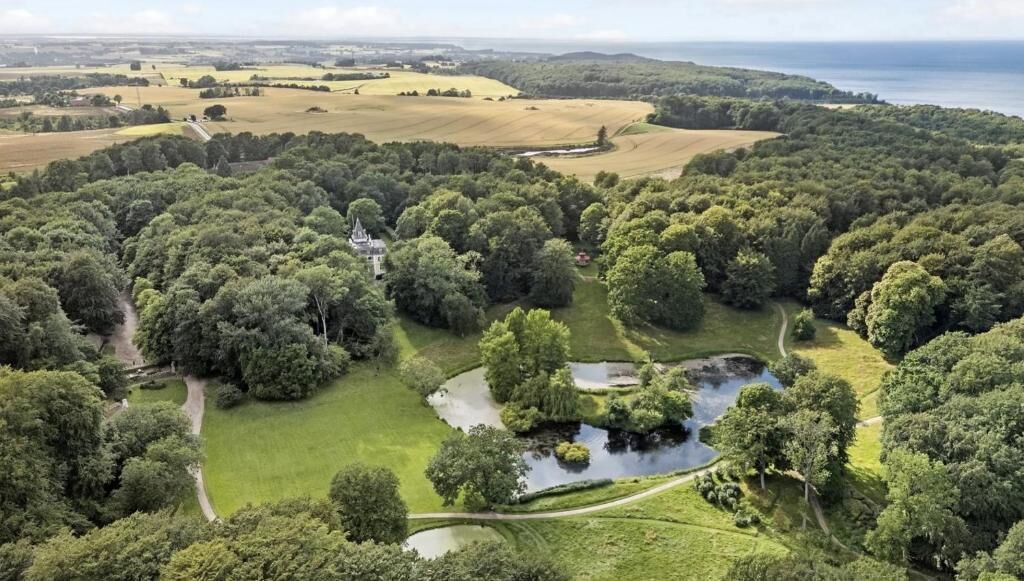 A lush landscape with a manor house surrounded by dense trees and a pond.