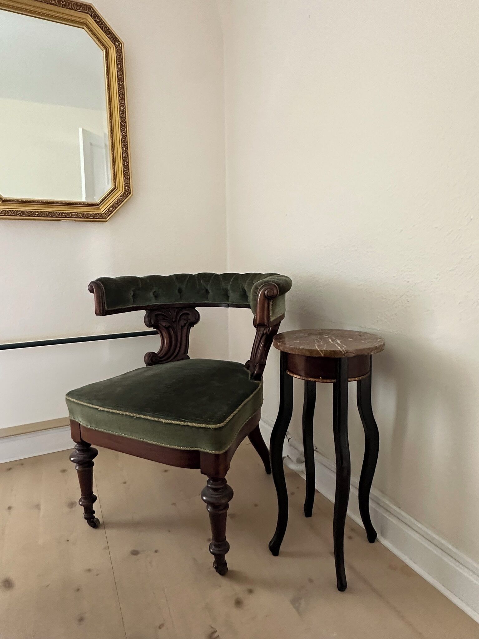A corner room with a green upholstered chair, a small round table, and a mirror on the wall.