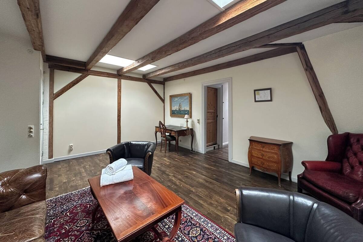 A room with a high ceiling, exposed wooden beams, a dining table with chairs, a red leather chair, a wooden cabinet, and a painting on the wall.