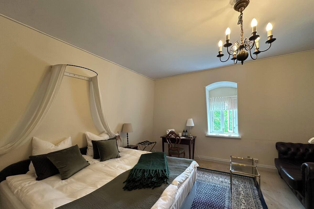A bedroom with a four-poster bed, a chandelier, a window with curtains, a desk with a lamp, and a leather armchair.