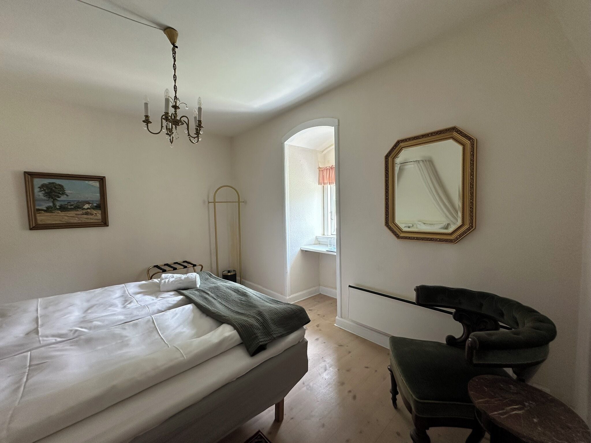 A bedroom with a bed, a chair, a mirror, a painting, and a chandelier.