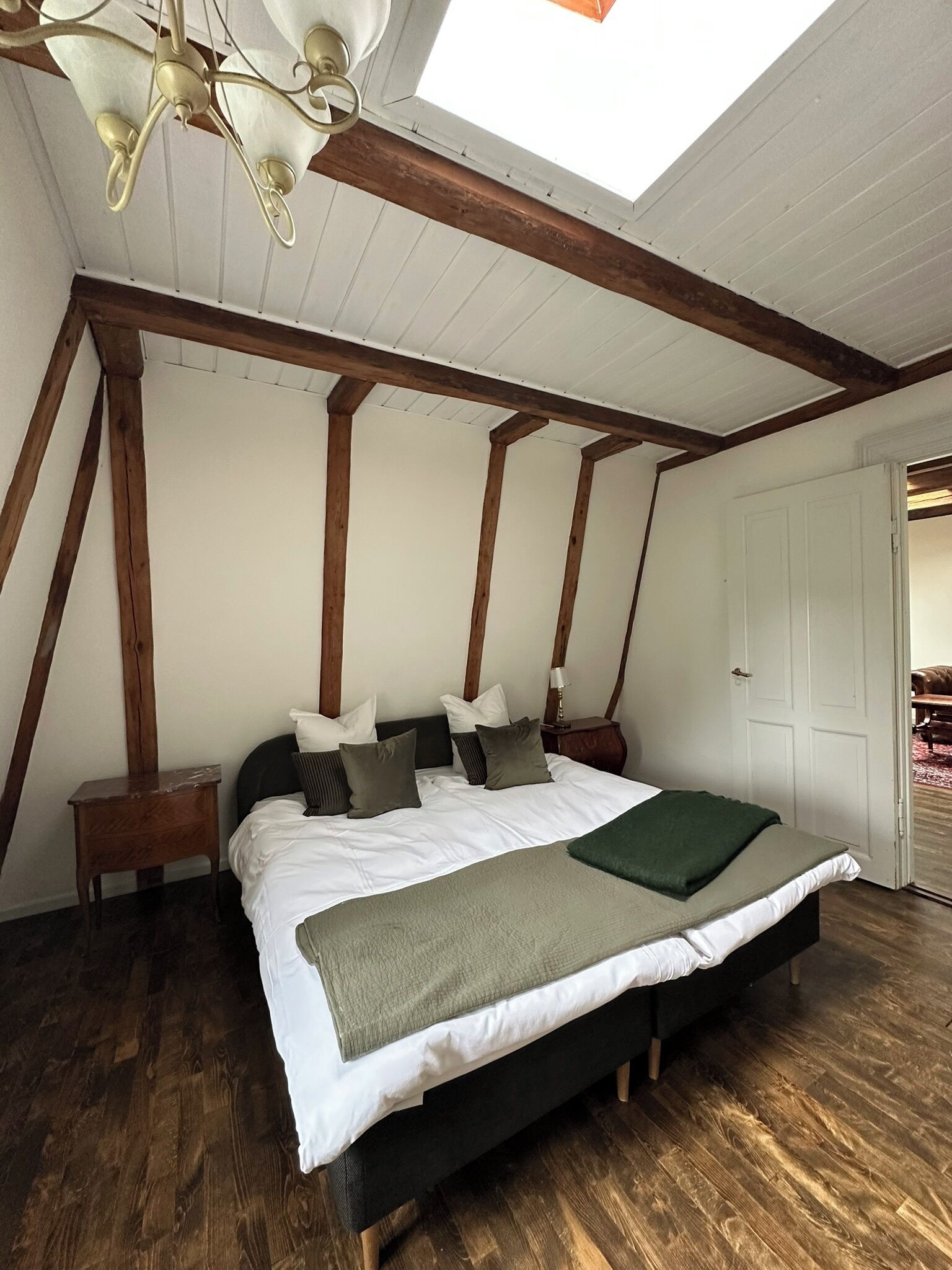Ein Schlafzimmer mit Oberlicht, Holzbalken, einem Bett mit Kissen, einem Nachttisch und einer Tür.