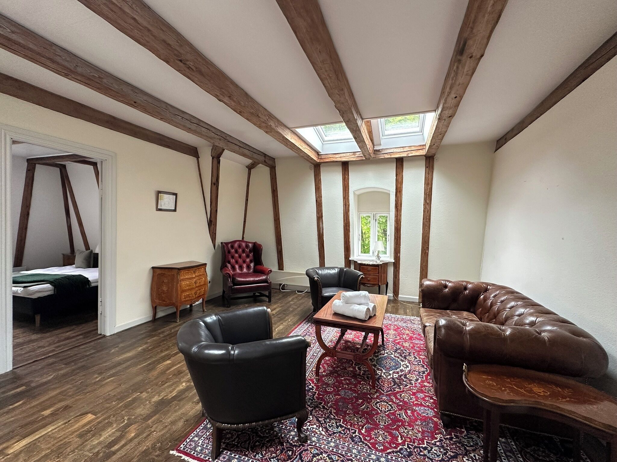 A room with a high ceiling, exposed wooden beams, a skylight, a red patterned rug, leather furniture, and a bed with a green blanket.