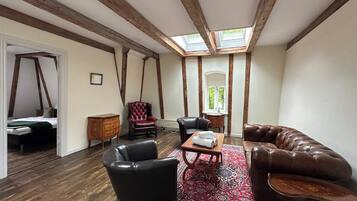 A room with a high ceiling, exposed wooden beams, a skylight, a red patterned rug, leather furniture, and a bed with a green blanket.