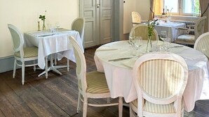 A dining room with two tables, chairs, a painting, and a window with curtains. - Liselund Ny Slot (Borre)