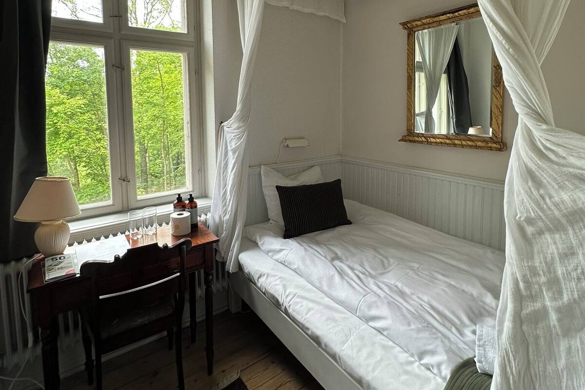 A bedroom with a single bed, a bedside table, a chair, a window with curtains, and a mirror.