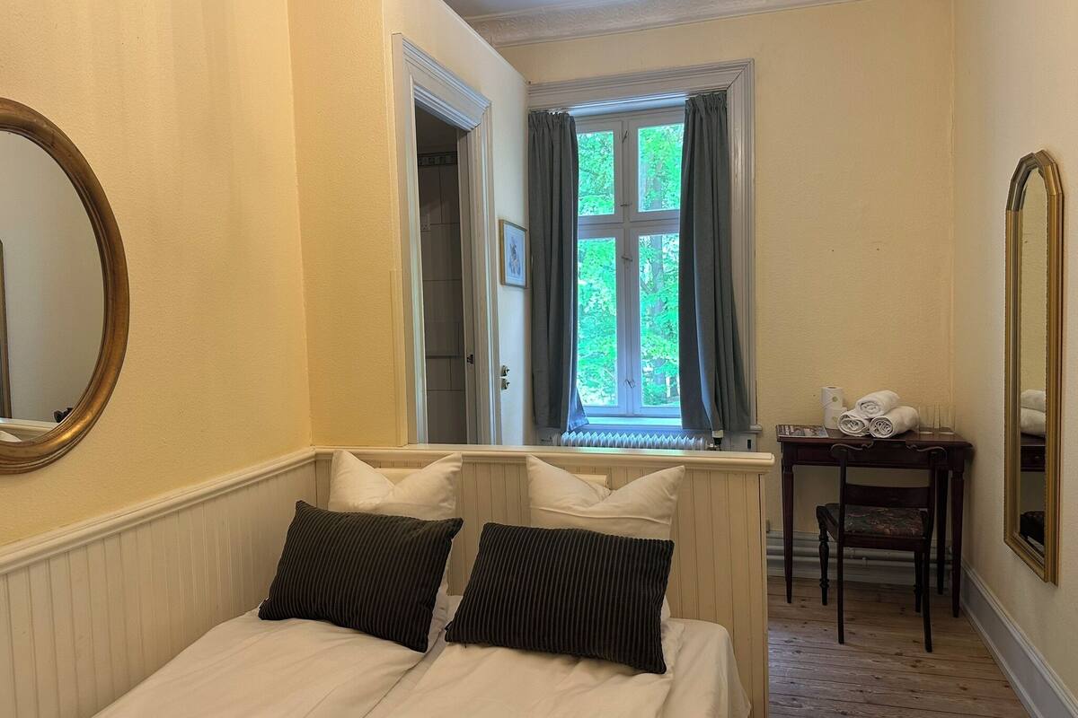 A bedroom with a bed, two pillows, a window with curtains, a chair, a small table with towels, and a mirror.