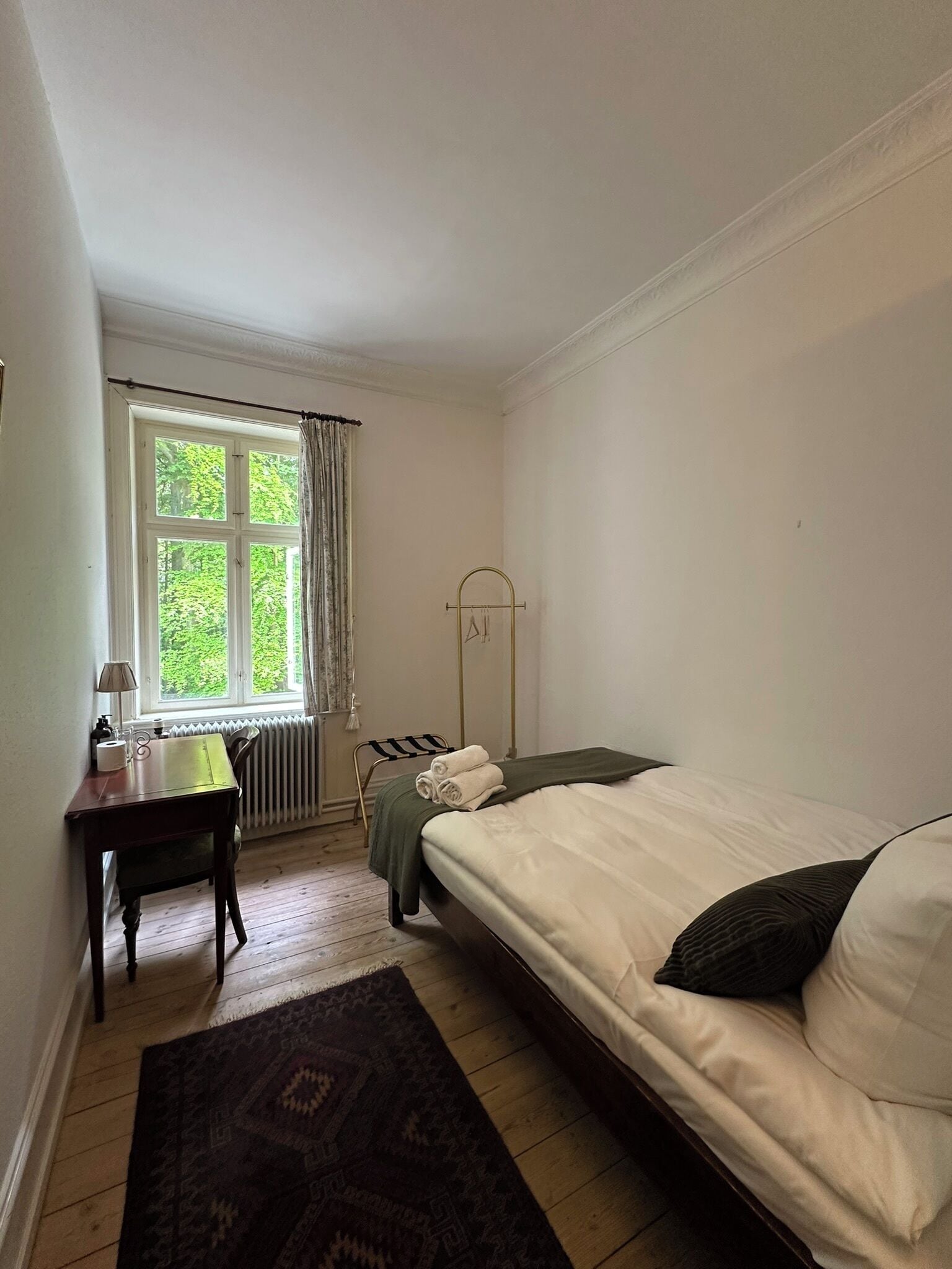 A bedroom with a bed, a desk, a radiator, and a window with curtains.