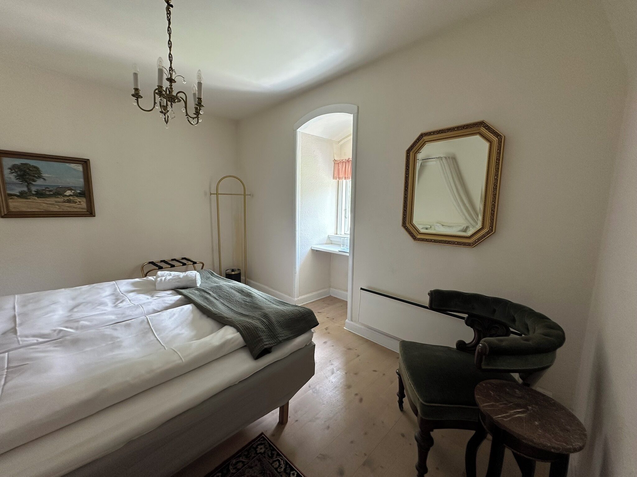 A bedroom with a bed, a chair, a mirror, and a painting on the wall.