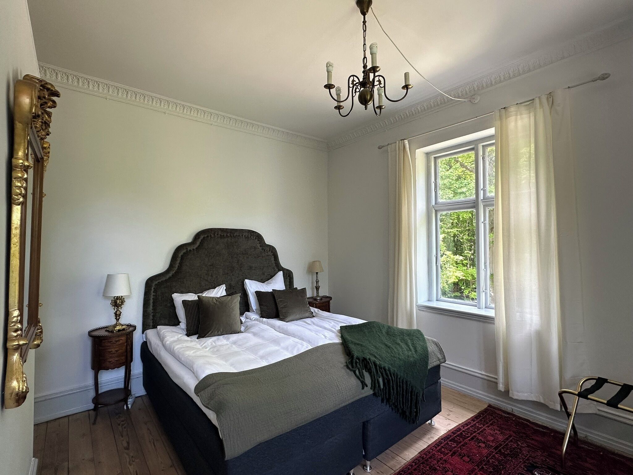 A bedroom with a large bed, a chandelier, a window with curtains, and a mirror on the wall.