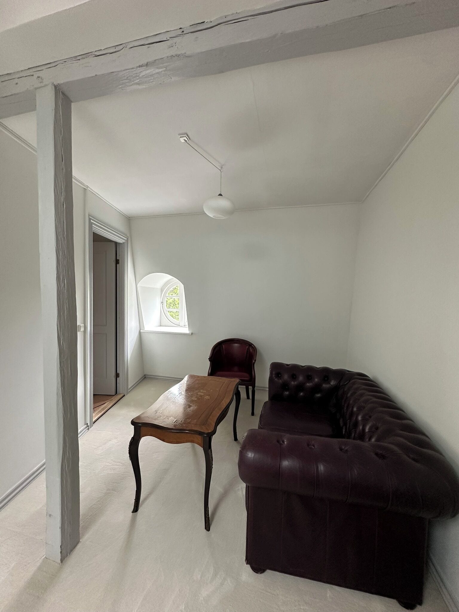A room with a dark leather sofa, a wooden coffee table, and a red leather chair. There is a window with a view of greenery outside.