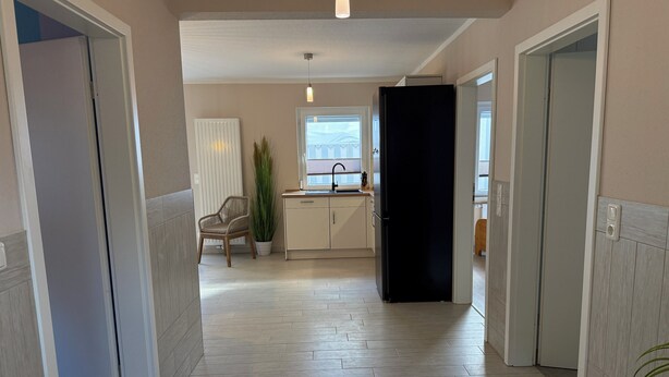 Un pasillo con una zona de cocina, una silla, una planta en maceta y una puerta negra.
