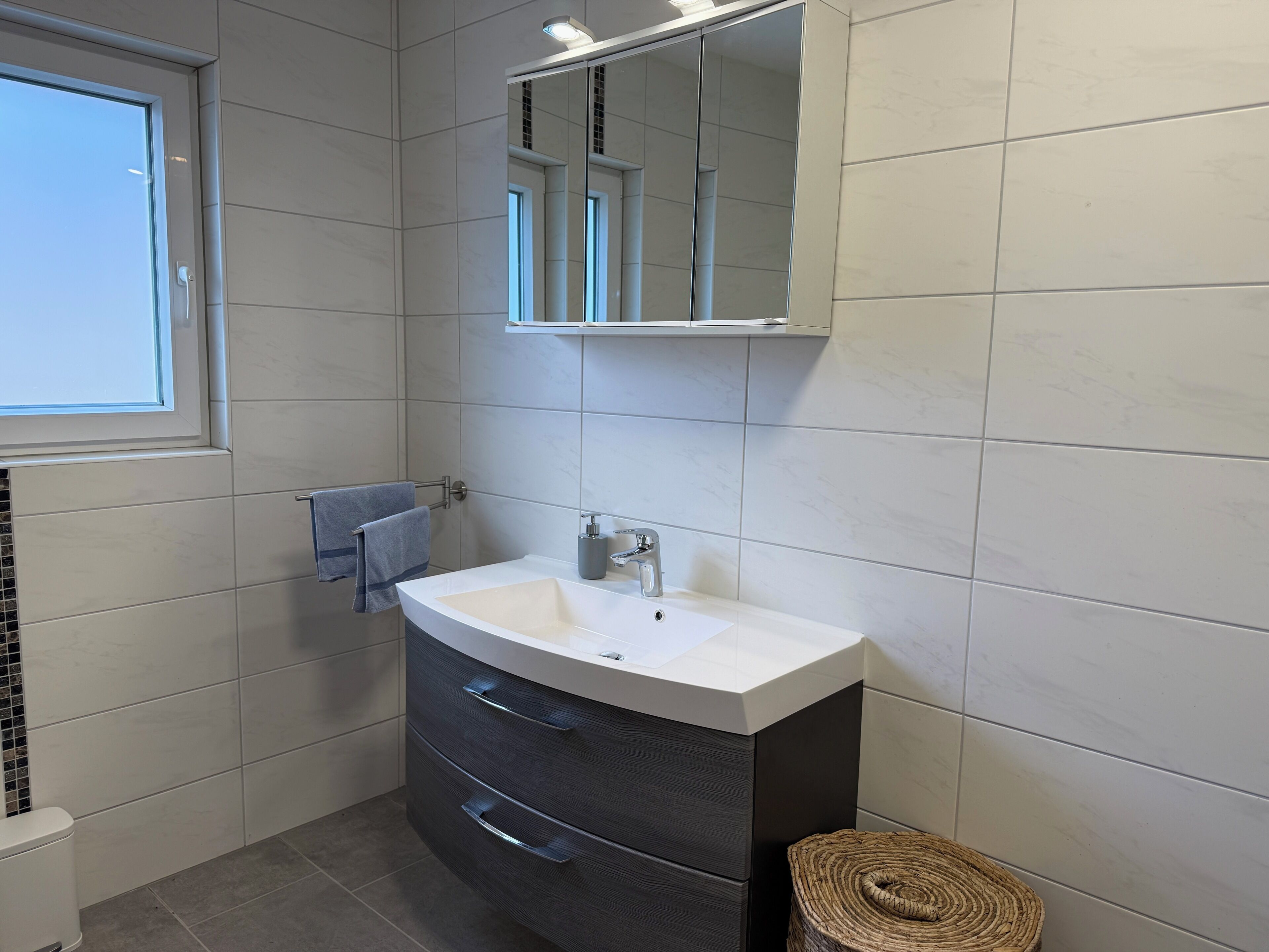 Ein modernes Badezimmer mit einem weißen Waschbecken, dunklen Schränken, einem Spiegel und einem Fenster.