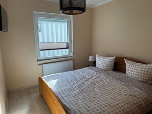 A bedroom with a bed, a window with blinds, a radiator, a bedside table with a lamp, and a ceiling light fixture. - Vacation home Pirat Störtebeker Dangast (Varel)