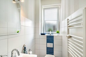 Een badkamer met een witte wastafel, een spiegel, een raam met zonwering, een handdoekhouder en een radiator.