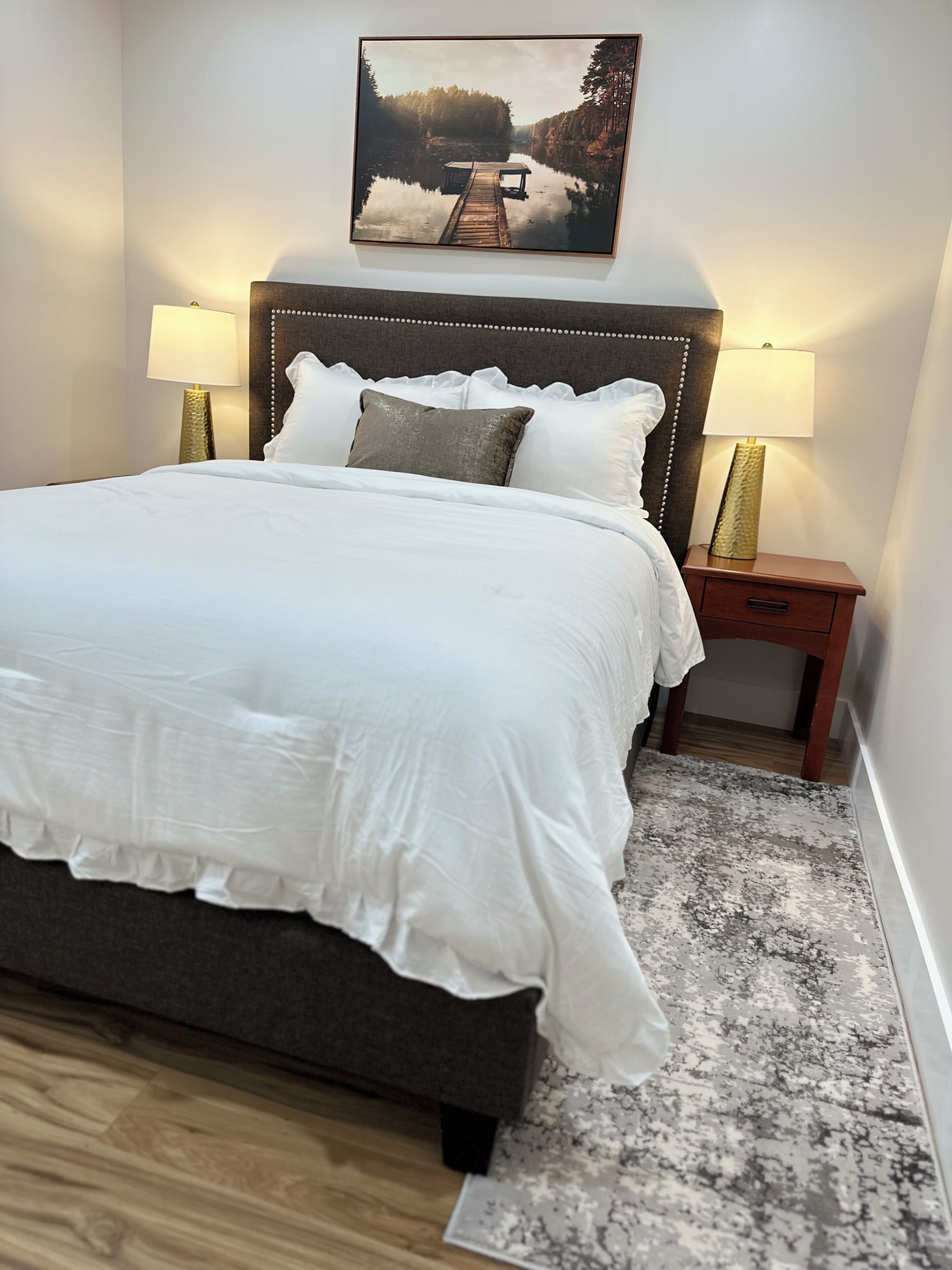 A neatly made bed with white bedding, a headboard with a decorative pattern, two bedside lamps, and a framed picture above the bed.