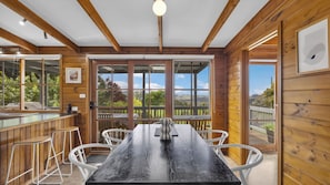 A wooden interior with a large table and chairs, a view through glass doors, and a wooden deck outside.