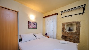 A bedroom with a bed, a wall-mounted shelf, a decorative wall piece, a window with curtains, and a stone wall feature. - One-Bedroom Apartment Senko A2 (Trpanj)