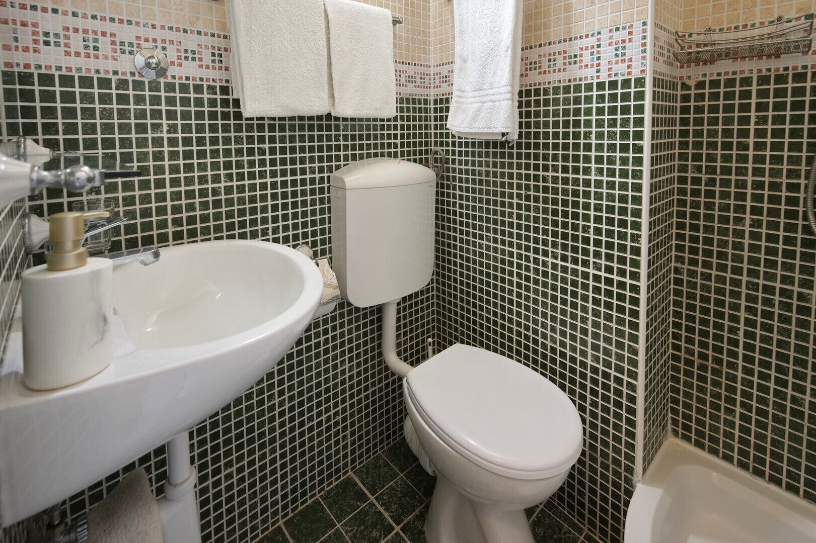 A bathroom with a white sink, toilet, and bathtub, tiled with green and white mosaic tiles.