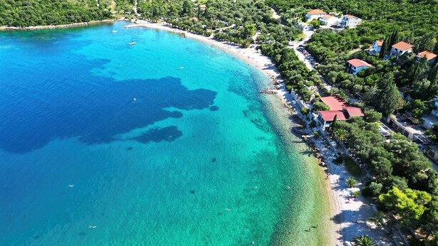 Vista aérea de una zona costera con aguas turquesas cristalinas, una playa de arena y una hilera de casas con tejados rojos.