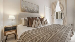 A bedroom with a bed, a nightstand, a lamp, a painting, and a window with curtains. - Hertford Flat 5 (London)