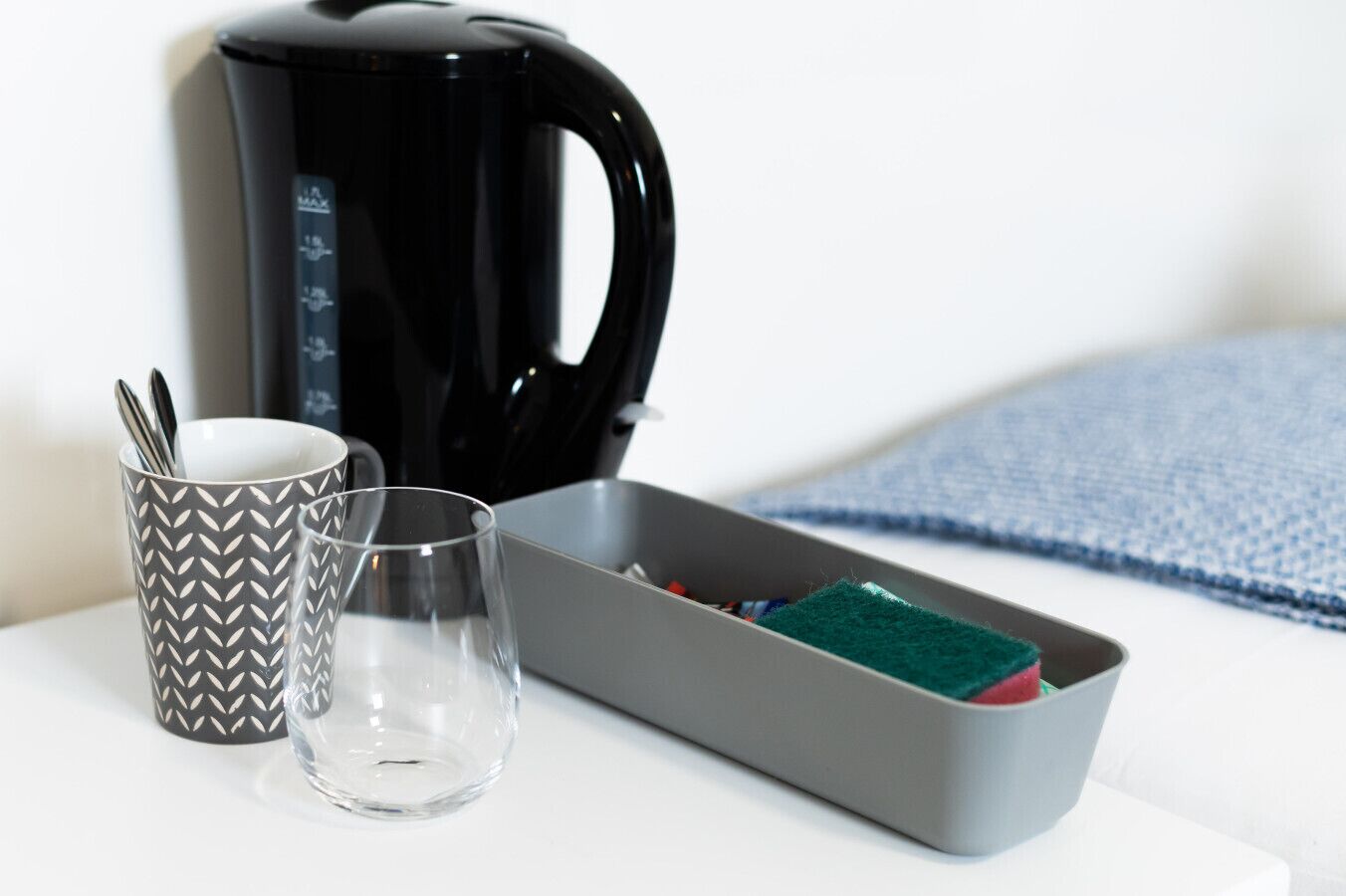 A black electric kettle, a patterned mug, a clear glass, and a grey box with a green sponge.