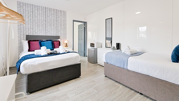 A modern hotel room with a large bed, a smaller bed, a desk, a mirror, a wardrobe, and a kitchenette. - Private En-Suite Studio with TV, Fridge & Free Parking (Saint Thomas)