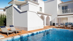 En moderne, hvid bygning med swimmingpool, liggestole og en overdækket patio.