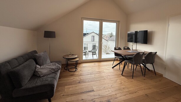 Un cuarto estilo loft con techo inclinado, un sofá, una mesa de comedor con sillas, un televisor y vistas al exterior.
