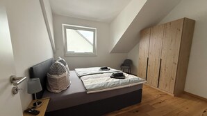 Un dormitorio con cama, ventana, armario y mesita de noche con lámpara y control remoto.