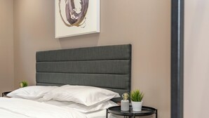 A neatly made bed with white linens, a grey upholstered headboard, and a small bedside table with a potted plant and a decorative item. - Morningside Private Residence (Edinburgh)