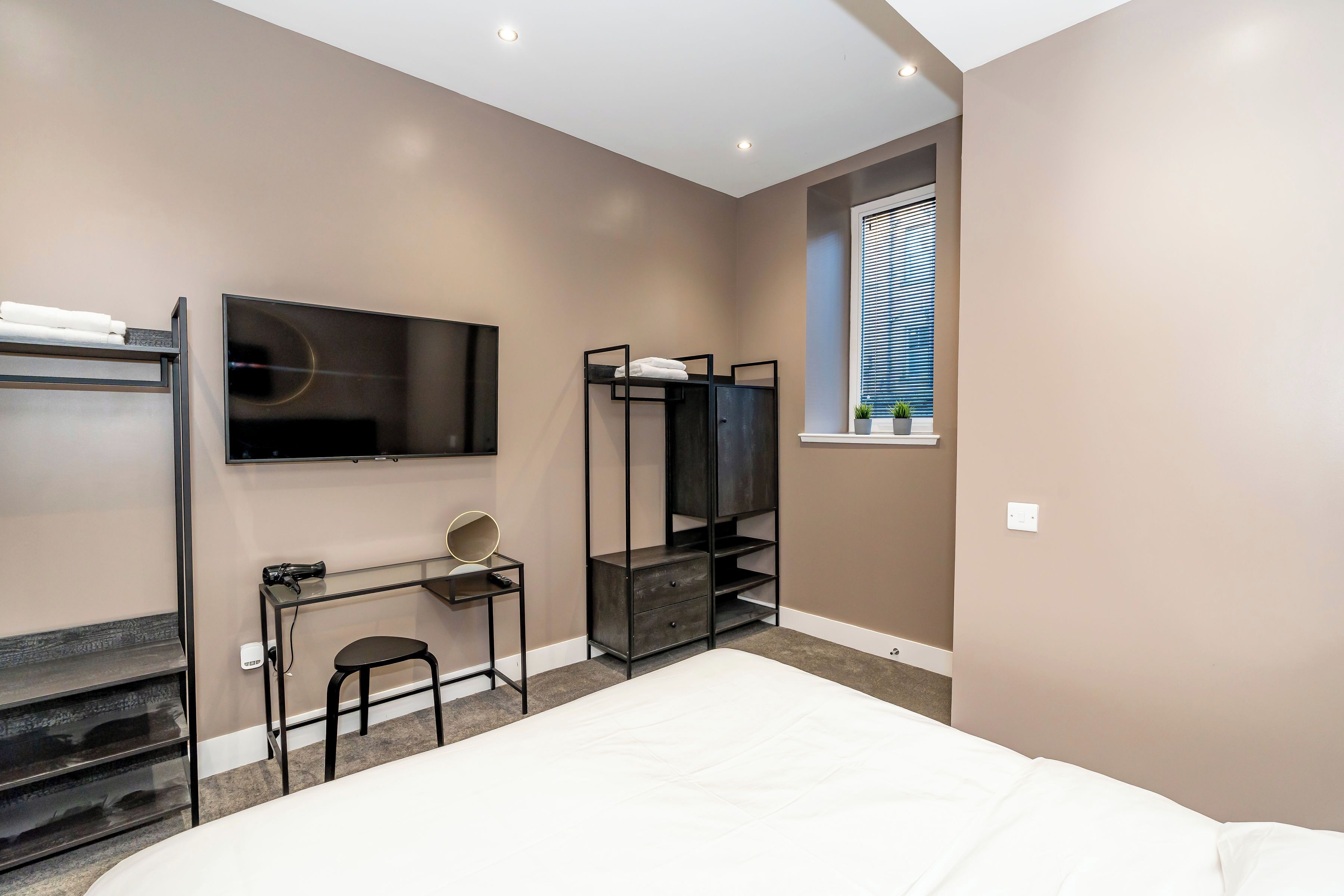 A bedroom with a bed, a TV mounted on the wall, a desk with a chair, a coat rack, and a window with blinds.