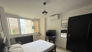 Un dormitorio con cama, escritorio, silla, ventilador, televisor y puerta.