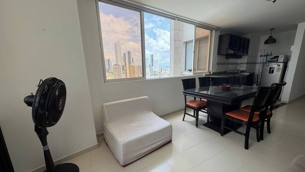 Una cocina moderna con comedor, un ventilador de pie, un sofá blanco y un ventanal con vista a la ciudad.