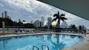 Una piscina con sillas de descanso, una palmera y un paisaje urbano al fondo.