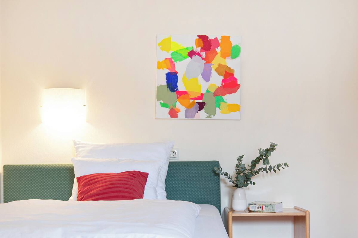 Una cama bien hecha con almohadas blancas y rojas, un cabecero verde, una mesita de noche con un jarrón de follaje y un cuadro abstracto y colorido en la pared.