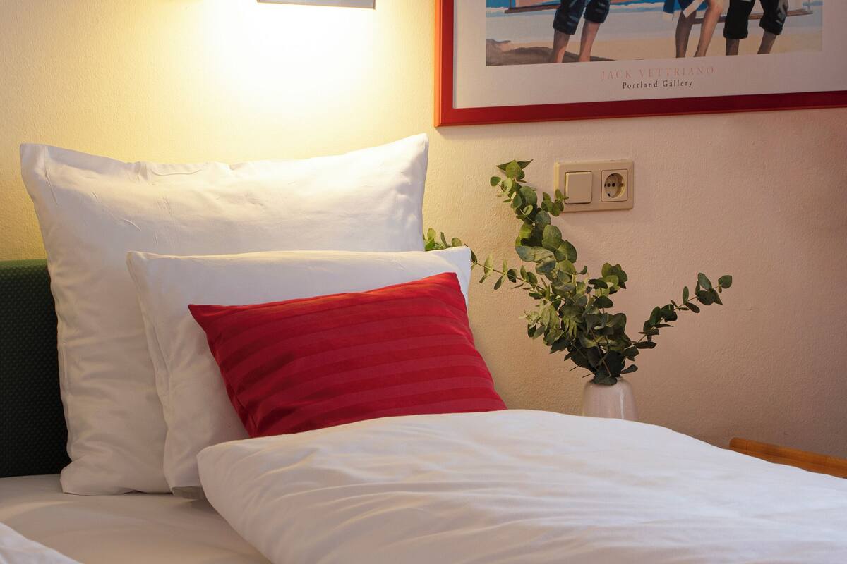 Una cama bien hecha con almohadas blancas y rojas, una lámpara de pared y un cuadro enmarcado encima.