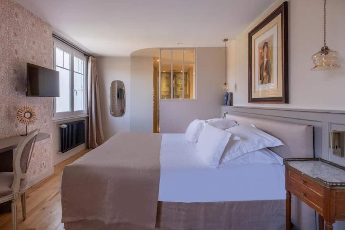 A bedroom with a bed, a desk, a chair, a TV, a framed picture, and a window.
